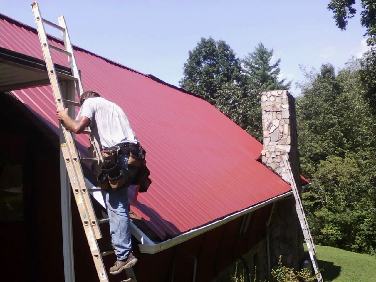 Roofer on a ladder beside a red metal roof and stone chimney — Soffit & Fascia Repair by Roofing Brothers in Riegelsville